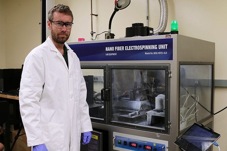 Army researcher in white lab coat in front of a Nano Fiber ElectroSpinning unit creating engineered carbon.