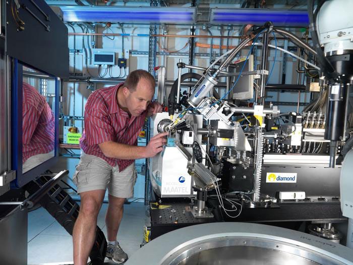 Male scientist wearing khaki shorts peers at large metal machines with lots of tubes and industrial piping in the background