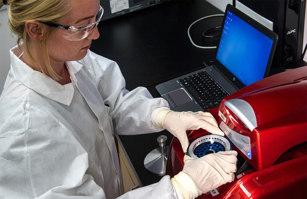 A woman wearing protective suit, gloves and goggles handles a round cylinder in a laboratory setting