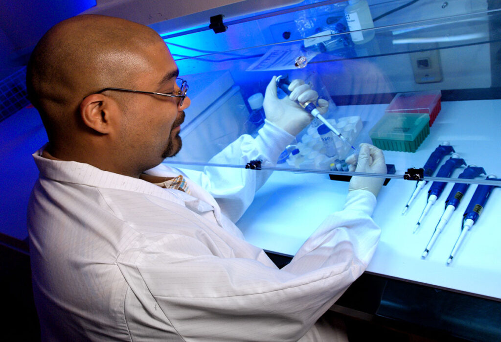 A laboratory scientist wearing a lab coat, safety glasses, and gloves is seated at a biosafety cabinet, carefully using a micropipette to transfer a liquid sample into a small vial. Inside the cabinet, various scientific tools and containers—such as pipette tips, sample tubes, and reagent racks—are neatly arranged. Multiple micropipettes are aligned on the work surface beside the scientist. The setting reflects a sterile, high-containment research environment typically used in genomics, infectious disease, or pharmaceutical research, underscoring the critical infrastructure at risk from NIH funding cuts.