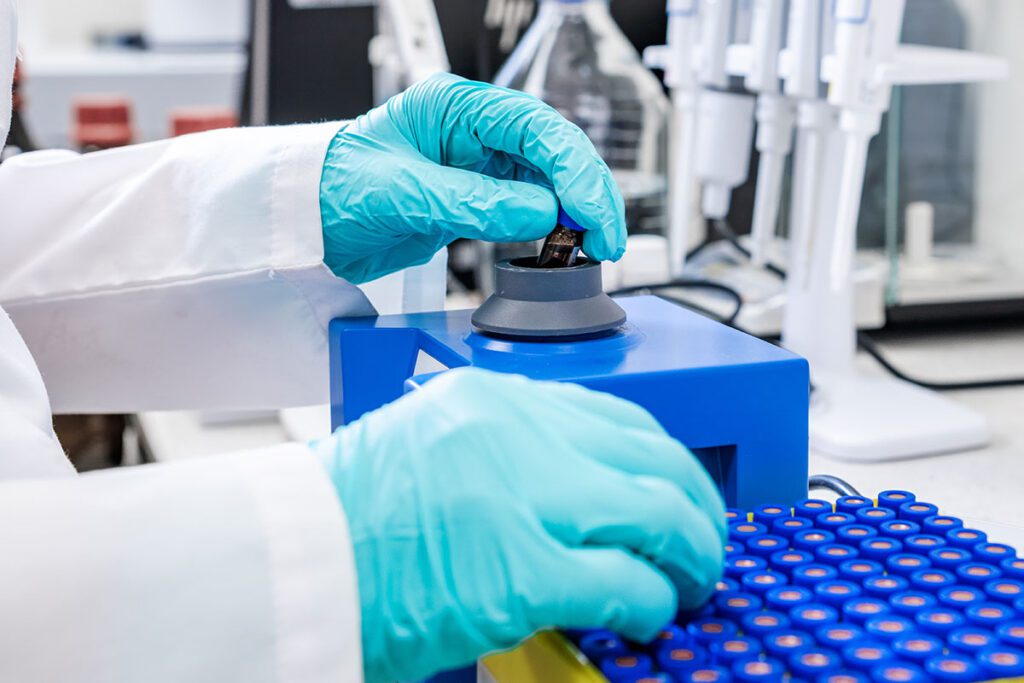 Gloved hands of a biomedical researcher placing a vial into a centrifuge in a laboratory, with rows of capped sample tubes and pipettes in the background, representing scientific research at risk due to proposed federal funding cuts.