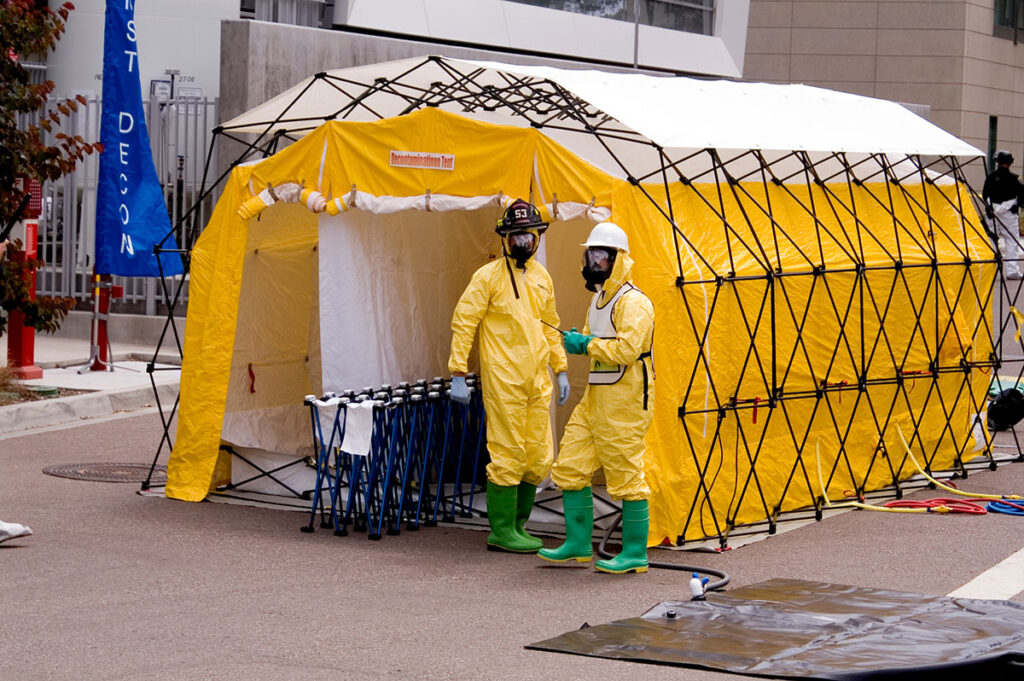 Two emergency responders in full-body protective suits and respiratory masks stand outside a portable decontamination tent used in chemical hazard response. The tent structure includes misting nozzles for decontamination, and nearby equipment includes folded support stands, hoses, and containment mats.