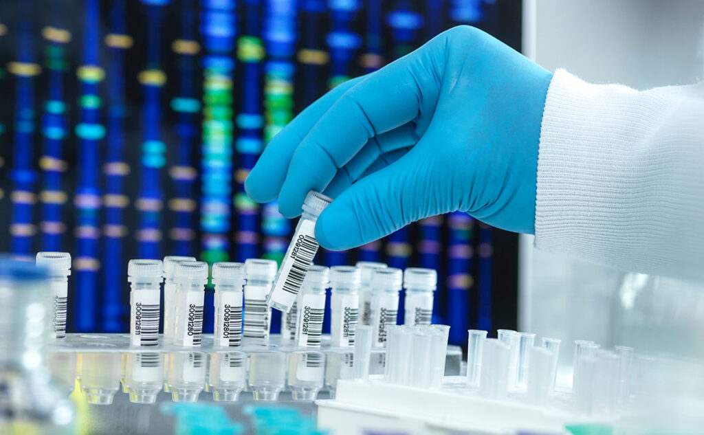 A gloved hand retrieves a labeled test vial from a rack of clinical samples in a laboratory setting. In the background, a digital display shows genetic sequencing patterns. The image conveys scientific research and clinical trial development related to mpox antiviral therapies.