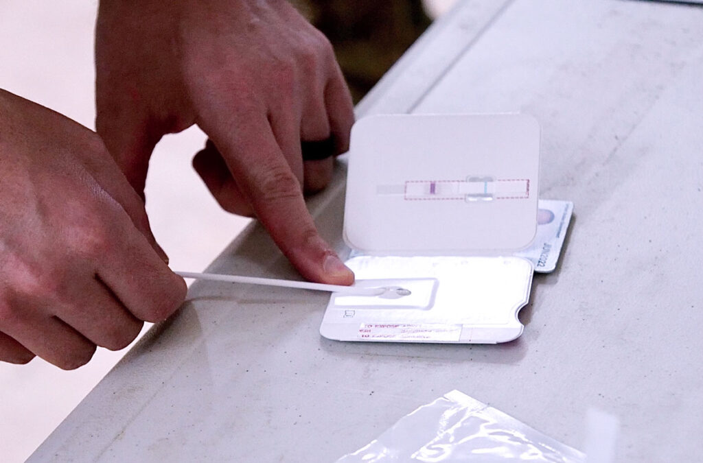 A person uses a cotton swab to place a nasal sample onto a COVID-19 rapid antigen test card positioned on a white table. The card includes a test strip and a control window to display results. This image illustrates the hands-on process of point-of-care SARS-CoV-2 diagnostic testing, highlighting the ease of use and quick turnaround time associated with rapid antigen tests during the pandemic.