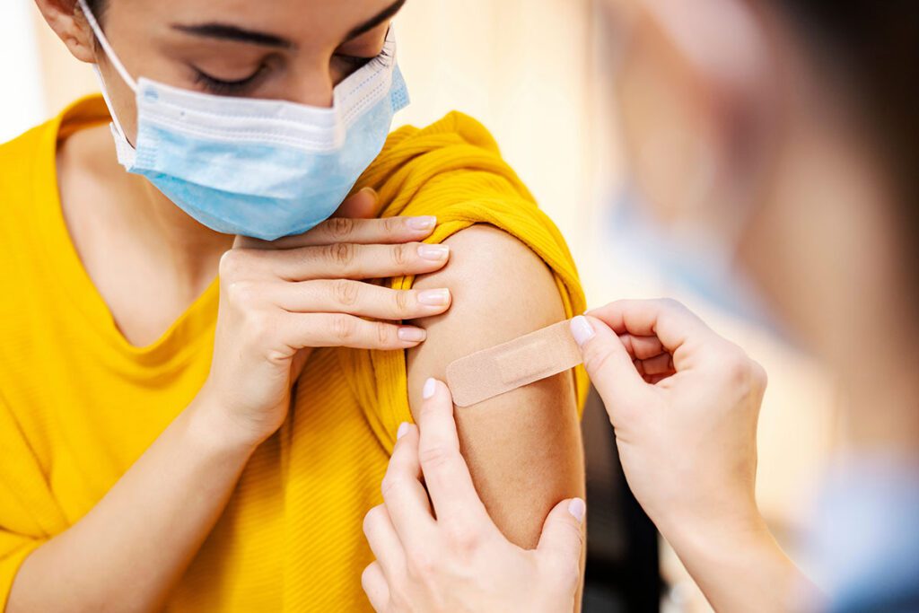 A close-up of a person receiving a vaccination. The individual is wearing a face mask and holding up their sleeve to expose their upper arm. A healthcare worker is carefully applying an adhesive bandage to the injection site. The scene takes place in a clinical setting, with the focus on the careful post-vaccine care process.
