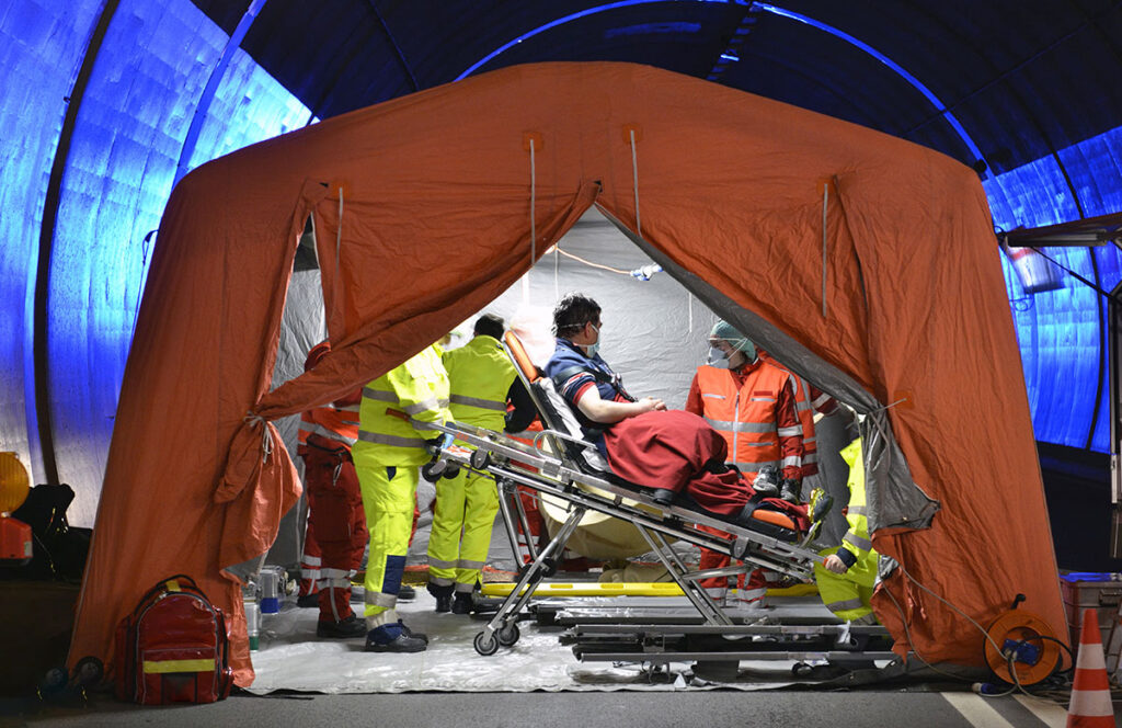 Emergency medical responders wearing protective gear attend to a patient on a stretcher inside a temporary triage tent set up in a tunnel. The scene illustrates a mass casualty preparedness drill focused on trauma care, infection control, and rapid response coordination.