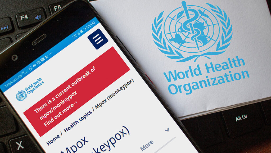 A smartphone screen shows the World Health Organization website with a banner warning about a current outbreak of mpox or monkeypox. Next to the phone is a document with the WHO logo. The image emphasizes WHO’s ongoing alert and response to the global mpox emergency.