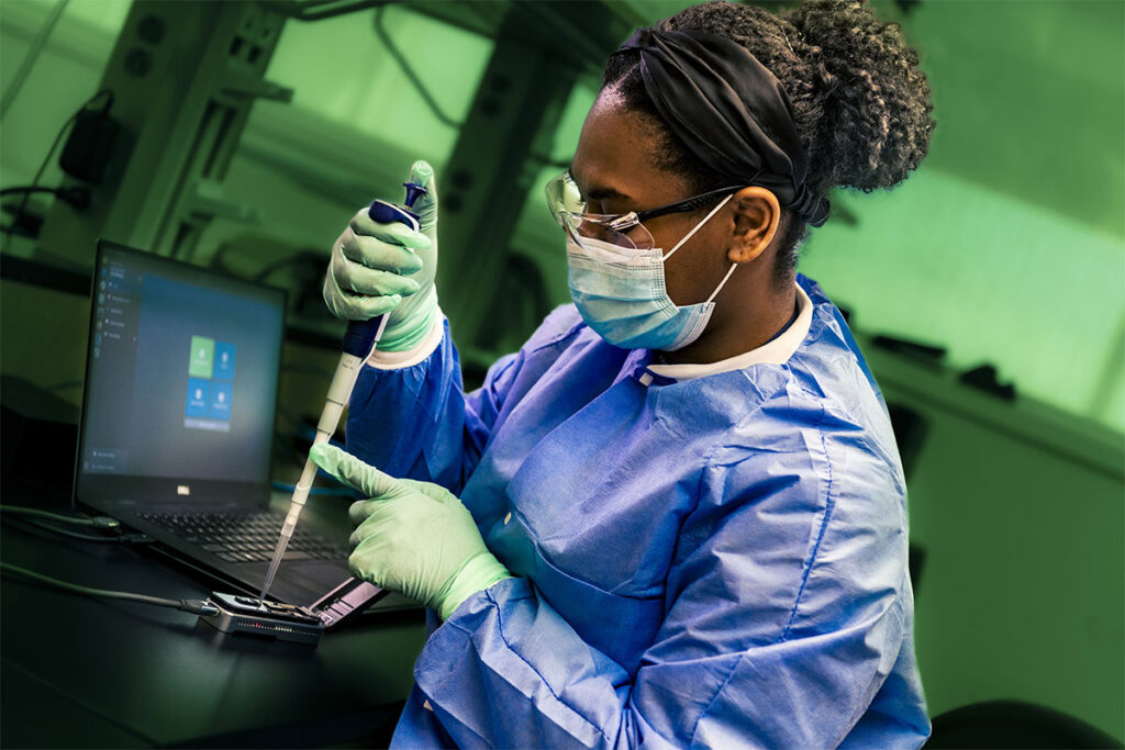 A public health laboratory scientist wearing a surgical mask, protective gown, and gloves carefully uses a pipette to load a sample into a portable genome sequencing device, which is connected to a laptop computer on a lab bench. The image illustrates the use of advanced molecular tools in infectious disease surveillance and outbreak response.