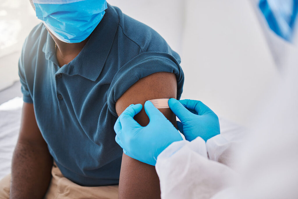 A person wearing a short-sleeved shirt receives a bandage on their upper arm from a gloved healthcare worker after receiving a vaccination. Both individuals are wearing face masks, and the setting appears to be a medical clinic.