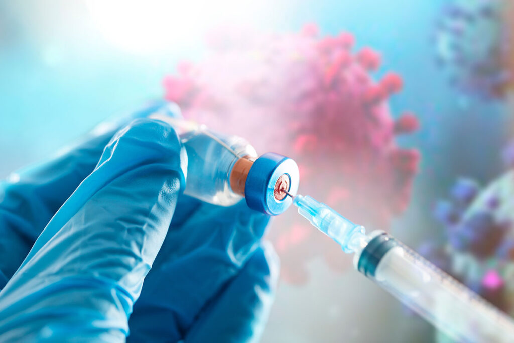 A gloved hand holds a vial of vaccine, while the opposite hand draws a dose into a syringe. The backdrop is a coronavirus drawing.