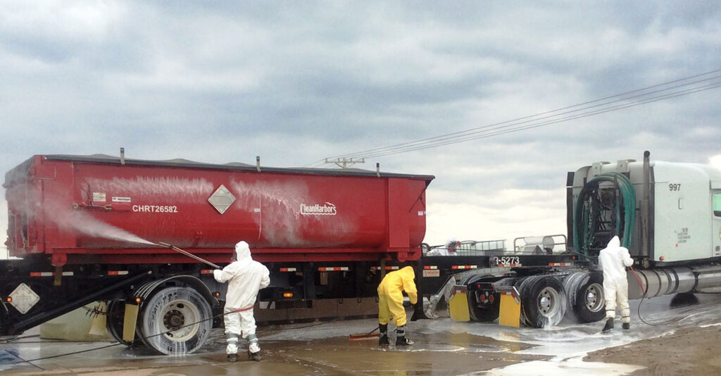Two workers in full-body protective white suits and one worker in a protective yellow suit conduct disinfection operations on a large red tanker truck. The workers use high-pressure water spraying equipment to wash the undercarriage and exterior of the vehicle on wet pavement. A tractor unit with a green cab is visible alongside the tanker. The scene takes place outdoors in overcast conditions with visible mist or spray from the cleaning operation.
