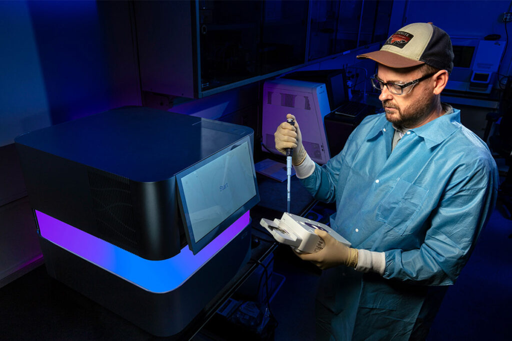 Pathogen-Agnostic Sequencing Finds Hidden Viruses A scientist wearing protective gloves and glasses works at a laboratory workstation, loading biological samples into an analytical instrument. The scientist holds a sample container in one hand while operating the equipment. The laboratory setting features sophisticated diagnostic equipment with display screens and the scientist is focused on the sample preparation and analysis process.