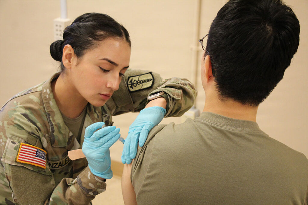 A military medical personnel wearing protective gloves administers an injection to a service member's arm. The provider, wearing military uniform with U.S. flag patch, focuses on the vaccination procedure. The service member faces away from the camera. The clinical setting shows a medical facility environment with basic equipment visible in the background.