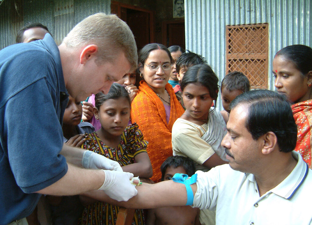 A public health worker wearing latex gloves draws a blood sample from the arm of a seated man, who has a tourniquet applied above the elbow. A group of adults and children stand close by, watching the procedure outside a rural structure with a corrugated metal wall. The setting conveys a field outbreak investigation in a village environment.