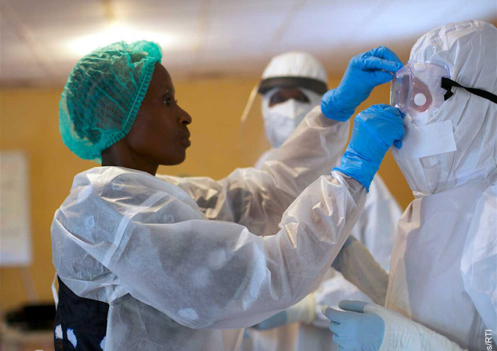 A health worker wearing protective gear demonstrates proper donning of a full protective suit to trainees. One trainee wears a head covering and protective gown while observing the demonstration. A second health worker in full protective equipment stands nearby. The training takes place in an indoor facility with yellow walls, showing hands-on instruction in outbreak response protocols.