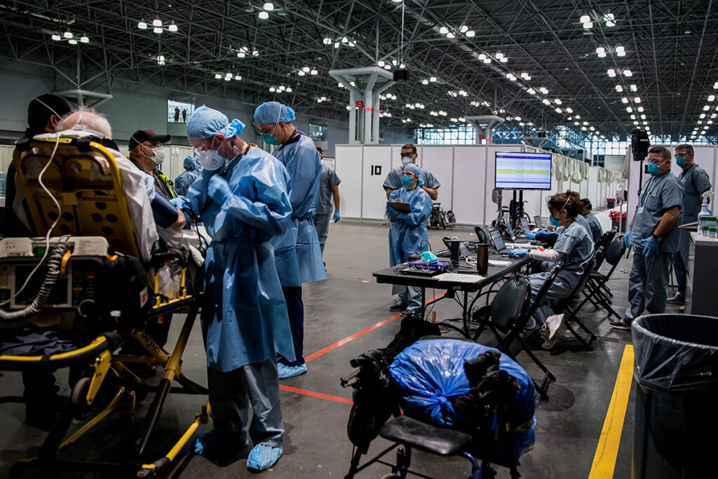 WHO Pandemic Agreement Stalls as Nations Fail to Resolve Pathogen-Sharing Deadlock A large convention center space has been converted into a field hospital with multiple patient care stations separated by temporary barriers. Medical personnel in protective equipment work at various stations throughout the vast open floor. Equipment including ventilators, monitoring devices, and medical supplies are visible. Modular workspace structures and organized care zones demonstrate how civilian infrastructure was repurposed for pandemic response operations during the surge period.