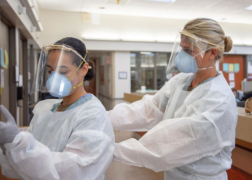 WHO Urged to Adopt Airborne Precautions as Default After Hantavirus Cruise Ship Outbreak One healthcare worker assists another in donning full protective equipment in a hospital corridor. The worker being fitted wears a light blue N95 respirator mask and clear face shield, while the assisting worker wears identical protective gear and white protective gown. Both are engaged in the fitting and adjustment process for the respiratory protection equipment. Clinical hallways and equipment are visible in the background.