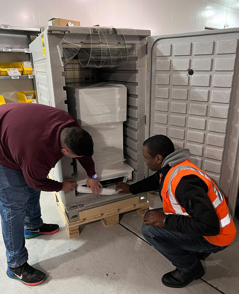 Two workers stand before a large insulated transport container with an open door. One worker in a safety vest crouches beside the container base while another leans forward to examine the interior. The container features thick insulation walls and internal space designed to hold temperature-sensitive medical supplies. Equipment for temperature monitoring and monitoring cables are visible at the top of the container.