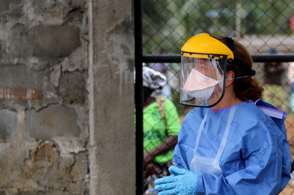 When Prevention Infrastructure Fails, Outbreaks Follow A healthcare worker wearing protective equipment stands at the entrance to a field hospital. The worker wears a yellow hard hat, clear face shield, blue protective gown, and gloves. Behind the worker, separated by fencing, other individuals are visible in the background. The temporary field hospital setting and surrounding area reflect the austere conditions of outbreak response operations.