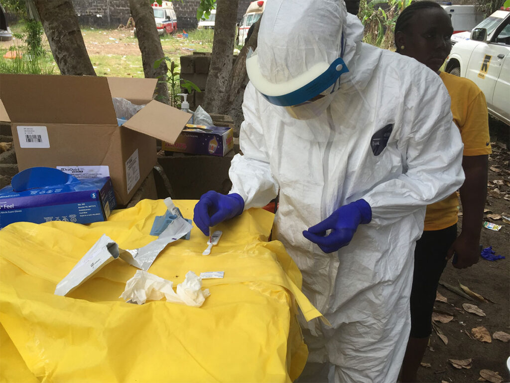 Ebola Strain Spreading in Congo and Uganda Has No Approved Vaccine A field epidemiologist wearing full protective equipment including a protective suit, face shield, and gloves performs rapid diagnostic testing at an outdoor work station. The epidemiologist holds testing materials and equipment on a work surface covered with protective yellow plastic. Cardboard boxes containing testing supplies and other field equipment are visible on the ground nearby. Community members can be seen in the background in a rural setting.