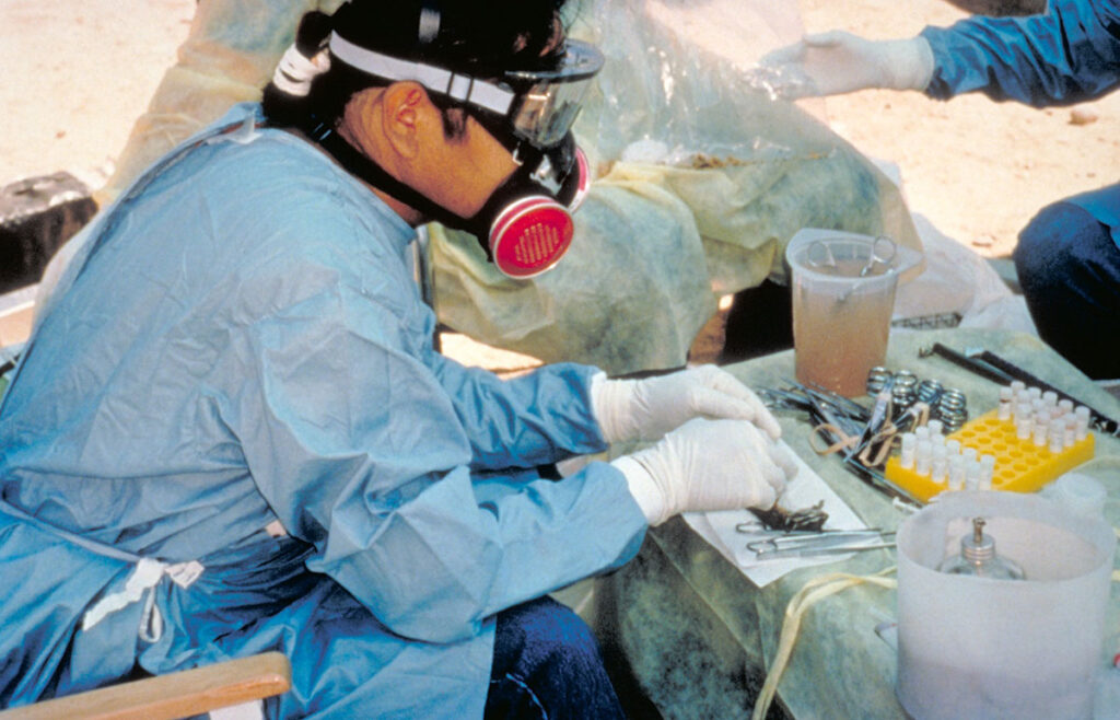 A field researcher wearing full protective equipment including a protective suit, respirator mask, and head covering sits outdoors while collecting tissue samples from trapped rodents. The researcher works at a collection station with various sampling tools, collection containers, and laboratory equipment visible on the work surface. Additional field equipment and another researcher can be seen in the background. Ready for the next image.