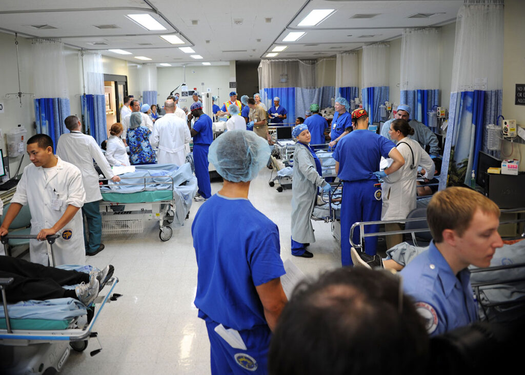 Inside the DHS Lab Developing Chemical Exposure Tools for Clinicians and First Responders A large hospital training space filled with medical personnel and multiple patient stretchers arranged throughout the room. Healthcare workers in scrubs and protective equipment move between stations, attending to simulated patients lying on beds and gurneys. Temporary partitions separate different treatment areas. The scene depicts a coordinated mass casualty exercise with numerous responders working simultaneously across the medical facility space.