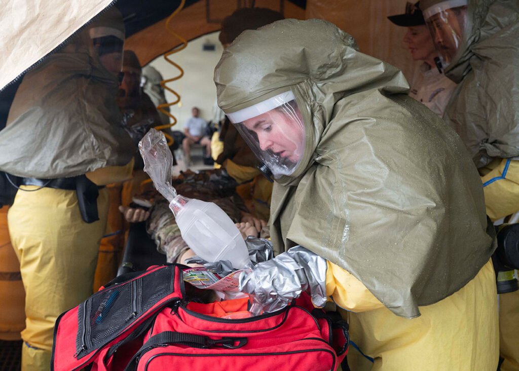 A medical responder wearing a full-face respirator and encapsulating protective suit leans over a patient on a stretcher inside a decontamination tent, preparing airway management supplies from a red medical bag. Additional personnel in protective suits are visible in the background.