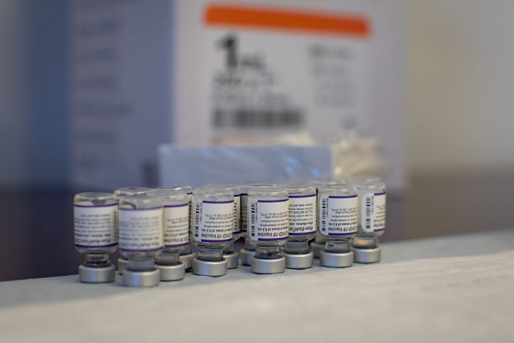 Multiple vaccine vials are arranged in a row on a white surface. The vials contain labeled vaccine material and are fitted with metal bases. Blurred equipment and packaging materials are visible in the background, showing a clinical preparation or storage area for vaccine distribution.