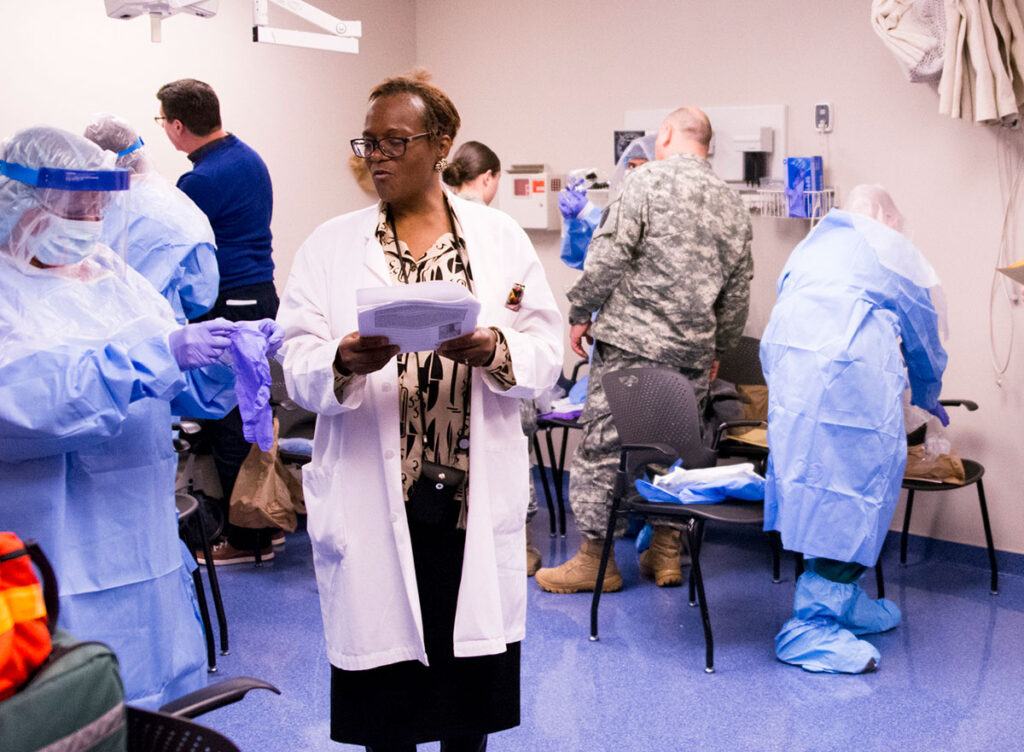 Nurses’ Union Warns Gutted CDC and Dismantled Global Health Infrastructure Leave U.S. Unprepared for Ebola, Hantavirus Threats Healthcare workers in a clinical training space practice donning and removing protective equipment. Multiple participants wear light blue protective gowns and gloves while working with partners. A trainer in a white coat with a clipboard observes and instructs the group. Equipment and supplies are visible on chairs and shelves throughout the room. The training demonstrates the buddy system approach to ensuring safe gear procedures.