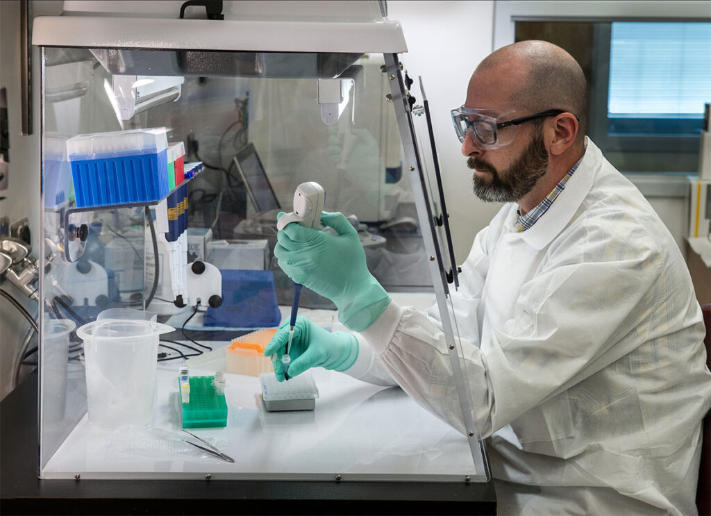 Federal Cuts to Public Health Infrastructure Threaten Hospital and Clinical Emergency Readiness A laboratory scientist wearing protective gloves and glasses works inside a biosafety cabinet, handling samples and equipment. The cabinet contains organized sample racks with multiple test tubes and laboratory materials. The scientist focuses on precise manipulation of samples at the work surface. Laboratory equipment and supplies are arranged within the contained workspace, typical of clinical pathogen screening operations.