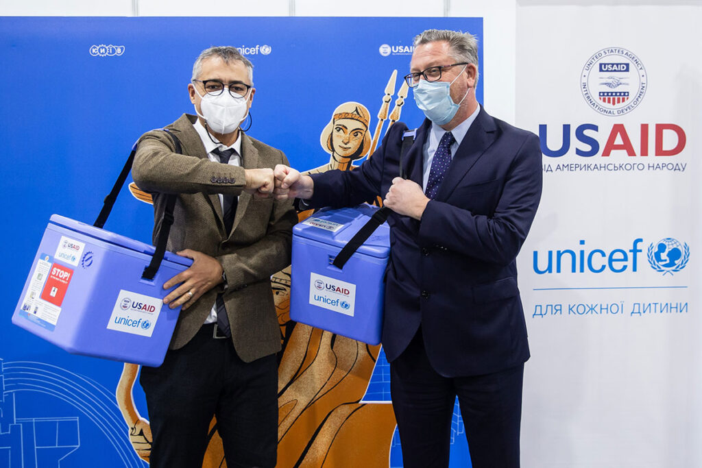 Two officials wearing face masks stand at a presentation event with a USAID/UNICEF backdrop. They exchange a fist bump while each holds an insulated cold chain container branded with UNICEF and USAID logos. A mascot figure is visible between them. The event setting shows organizational logos and text in multiple languages, indicating international health collaboration.