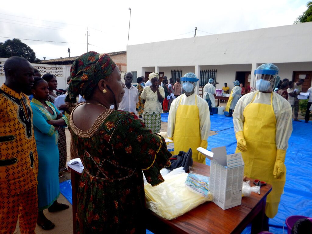 Before the First Case: Why Ebola Preparedness Must Move Upstream Healthcare workers participate in a community training session on infection prevention and personal protective equipment use. Two trainers wearing full protective suits including respirators and gowns demonstrate proper PPE donning and procedures to local healthcare workers and community members gathered around a table with educational materials. The training takes place in an outdoor facility setting with multiple participants observing the practical demonstration.