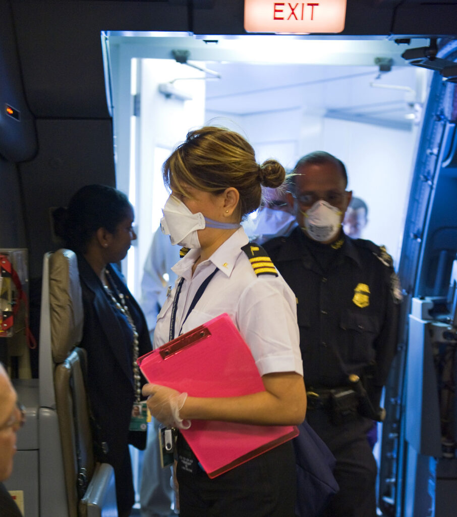 A health official in a white pilot-style uniform and face mask stands inside an aircraft doorway holding a clipboard. The official is positioned near the aircraft exit sign and surrounded by other uniformed personnel and passengers. The scene depicts an in-flight health screening or assessment operation, with the aircraft interior visible and multiple individuals present in the confined space.