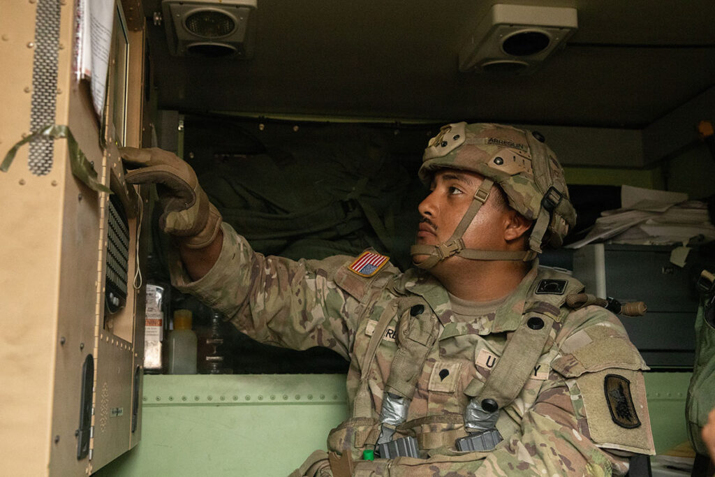 A military specialist in camouflage uniform operates a mounted biological detection system during a training demonstration. The soldier reaches toward detection equipment installed on what appears to be a vehicle or portable platform. The operator is focused on the display or control interface of the detection device. Military equipment and structural elements of the training facility are visible in the background.