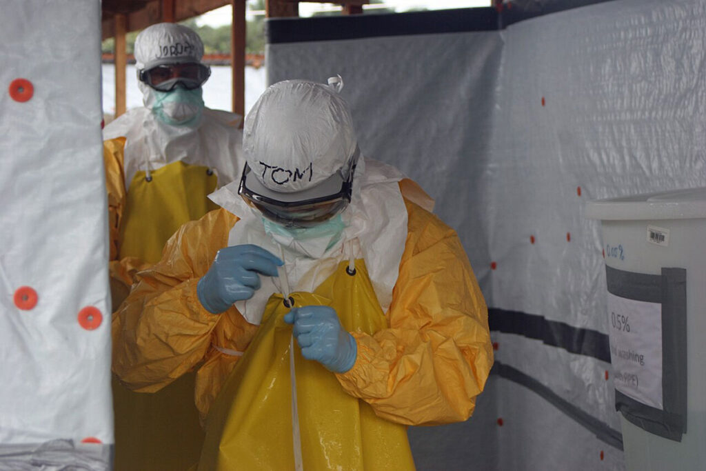 A person in full protective gear including a yellow outer suit, respirator, and goggles carefully removes protective equipment with assistance from another worker. The individual is positioned at the entrance to a containment structure with white plastic sheeting and marked decontamination zones. A staff member stands in the background providing decontamination support. Red marker indicators are visible on the plastic sheeting marking specific decontamination stations and contact points.