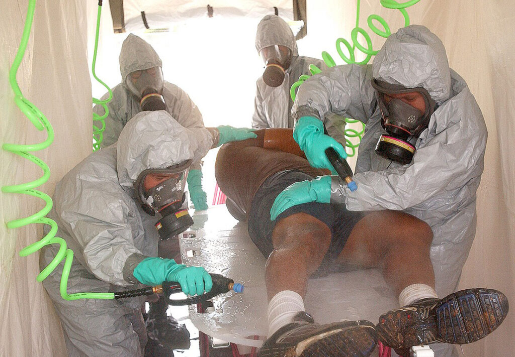 Multiple hospital personnel wearing full protective suits, respirators, and gloves conduct decontamination procedures on a simulated patient. The personnel work systematically around the patient using spray equipment and water delivery systems visible through flexible tubing. The clinical decontamination area shows the contained, methodical approach to patient decontamination in a hospital setting. Medical staff maintain coordinated positions to ensure thorough treatment.