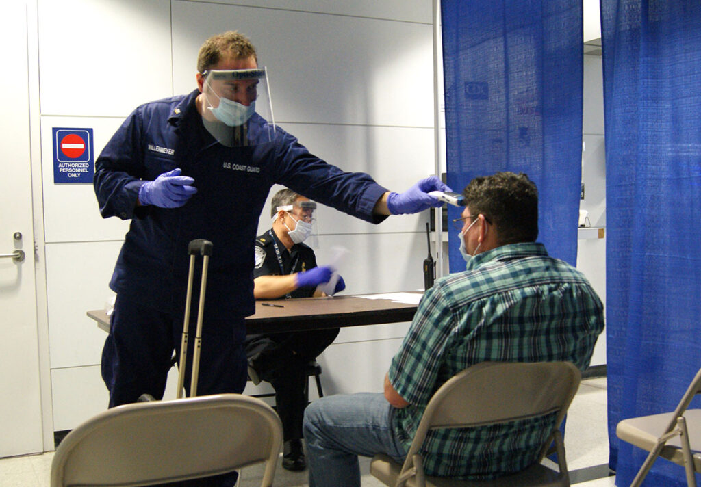 A U.S. Coast Guard officer wearing protective equipment including a face mask and gloves conducts a health screening of a passenger at an airport checkpoint. The officer stands behind a desk speaking with the traveler, who is seated. A second uniformed official is visible at the desk. An "Authorized Personnel Only" sign and screening partition are visible in the background, indicating an official border health screening area.