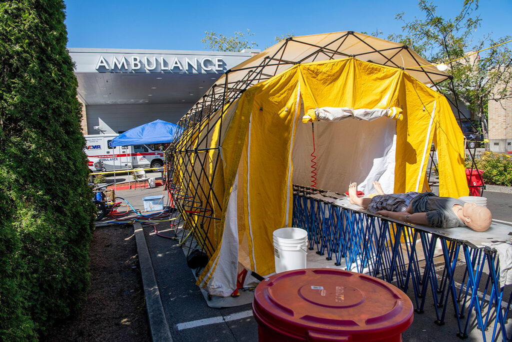 An outdoor decontamination tent set up beside a hospital ambulance entrance features a metal frame structure with fabric enclosure. A training mannequin lies on a stretcher inside the tent. Buckets, washing equipment, and containment basins are positioned for decontamination procedures. Ambulances and the hospital building are visible in the background, showing the medical facility context for the training exercise.