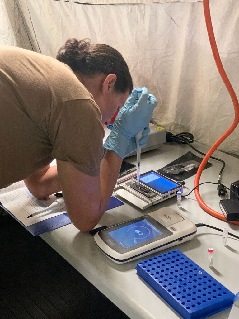 A microbiologist wearing protective gloves bends over a portable laboratory workstation set up inside a tent. The operator is loading or preparing samples using several connected instruments with display screens. Multiple pieces of equipment are visible on the white work surface, including devices showing data on screens, sample preparation trays with multiple wells, test tubes, and various cables and connections. Documentation and materials are visible at the workspace edge.