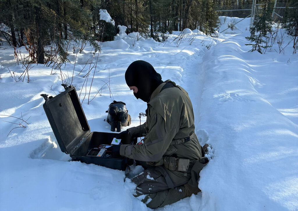 A military operator wearing cold-weather gear and a face covering kneels in deep snow while operating a portable sequencing device. The equipment consists of a ruggedized case with an open panel and connected scientific instruments. The operator is positioned in front of a snow-covered landscape with evergreen trees and dense forest in the background. A chain-link fence is visible through the trees.