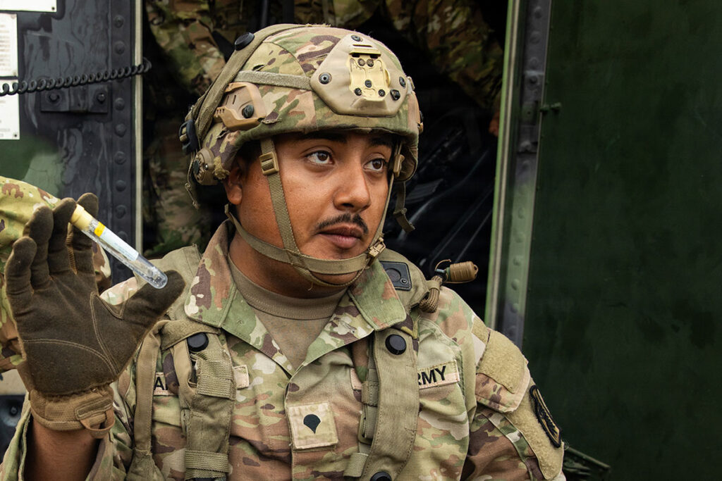 A soldier in camouflage uniform and combat helmet holds a handheld detection device. The portable equipment features a cylindrical sensor module and detection cartridge. The soldier is photographed in profile, demonstrating the compact size and field-portable design of the biological detection system. Military equipment and structural elements are visible in the background.