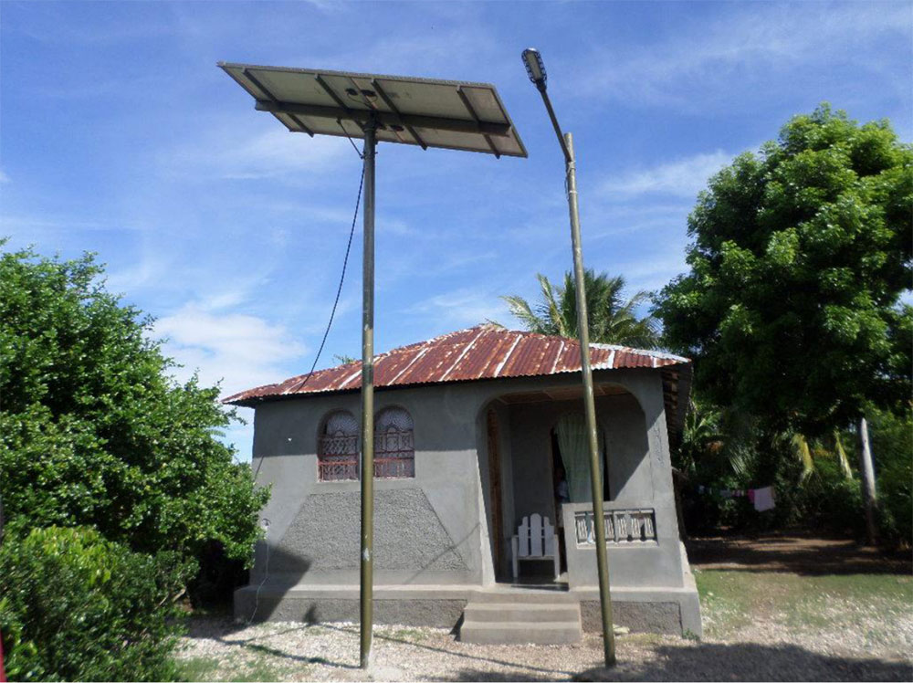 A small concrete outpost building with a corrugated metal roof stands in a rural tropical setting. Solar panels are mounted on tall poles beside the structure, providing power for refrigeration equipment inside. The building features two wooden doors and is surrounded by vegetation and trees. An electrical pole is visible nearby, showing the power infrastructure connecting the solar installation to the facility.