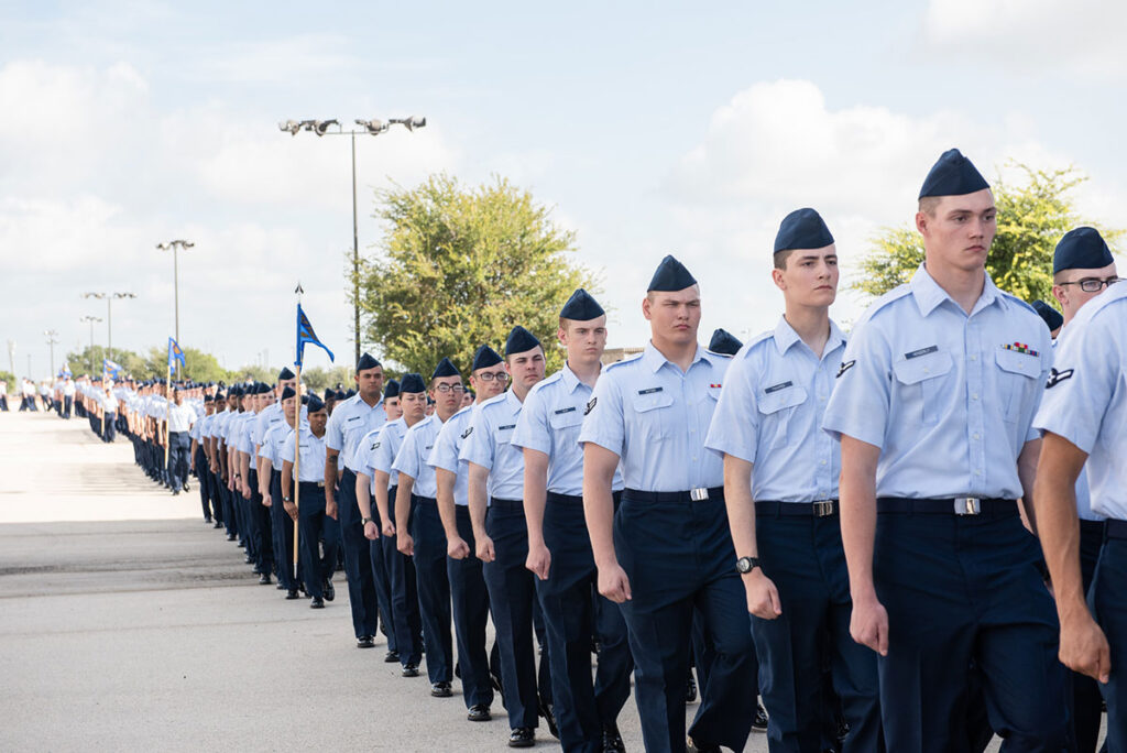 Military Reverses Flu Vaccine Mandate After Outbreak Sickens Hundreds at Air Force Base Hundreds of military trainees stand in formation during a graduation ceremony. Personnel are arranged in multiple rows, all wearing dress blues. Military flags and command personnel are visible overseeing the ceremony.