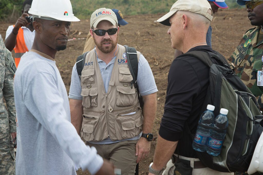 U.S. Dismantling of USAID Is Undermining Ebola Response, Experts Warn A USAID military liaison and project manager stand on a construction site with local personnel and military staff. The group discusses plans for the treatment facility on barren ground with construction materials visible. A construction worker wears a hard hat, and multiple individuals in work attire confer on the site. The scene depicts collaborative international outbreak response infrastructure development.