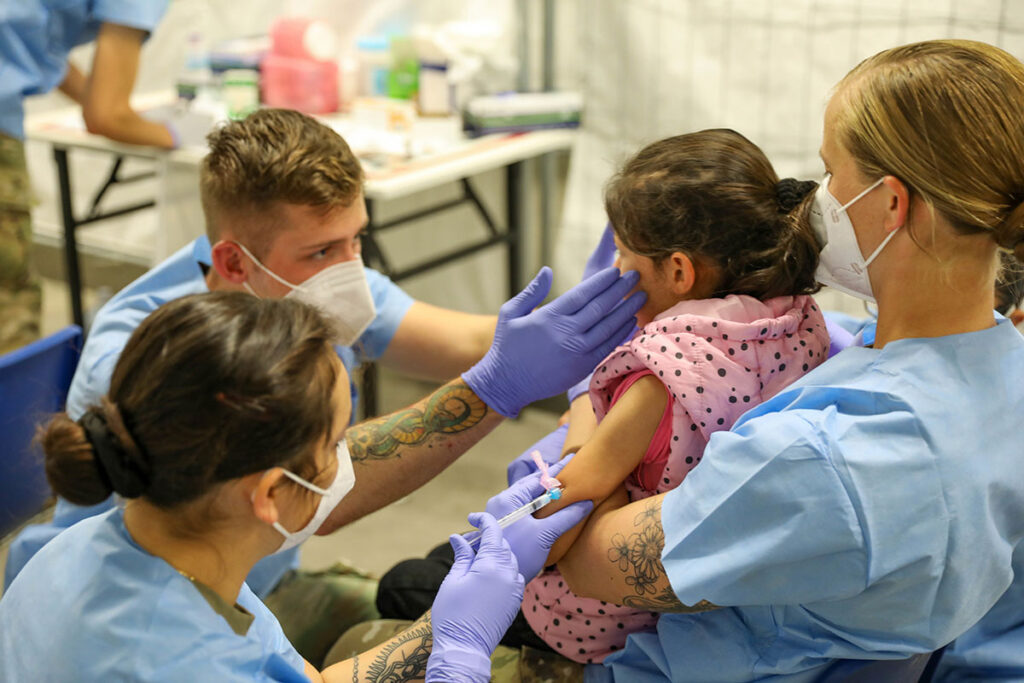Largest Study Yet Finds No Link Between MMR Timing and Autism A young child in a pink polka-dot jacket sits on an adult's lap while a healthcare worker in gloves and a mask administers an injection into her upper arm. Another healthcare worker steadies her chin, and a third person looks on nearby. All the adults wear surgical masks and protective gowns, and medical supplies are visible on a table in the background inside a tented facility.