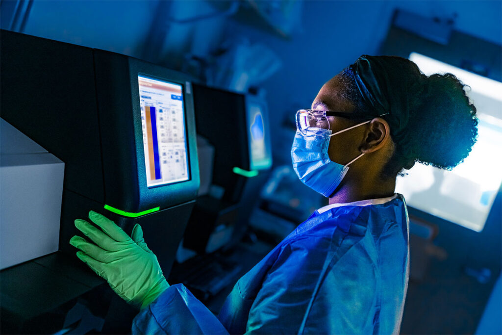 A laboratory scientist wearing protective gloves and a face mask works at a genomic sequencing workstation. The researcher is positioned at a computer terminal displaying data analysis software and charts on the screen. Laboratory equipment is visible in the background, including additional analytical instruments. The workspace shows typical modern research laboratory setup with specialized equipment for pathogen identification and genetic analysis.