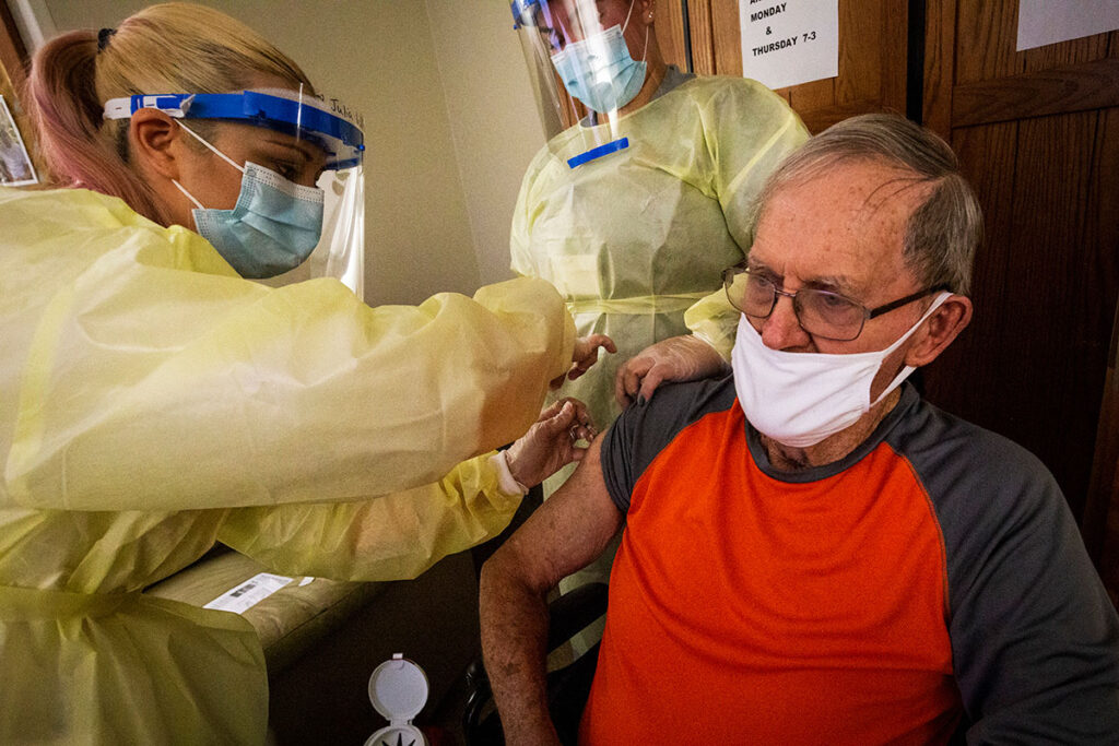 Updated COVID Vaccine Still Cuts Heart Risks — Especially for Older Adults, Study Finds A healthcare worker in full protective equipment administers a vaccine injection to an elderly male patient seated in a clinical setting. The provider wears a protective gown, face mask, and clear face shield. The patient wears a face mask and glasses. A schedule posted on the wall in the background indicates vaccination appointment times. The scene depicts a routine immunization procedure at a medical facility.