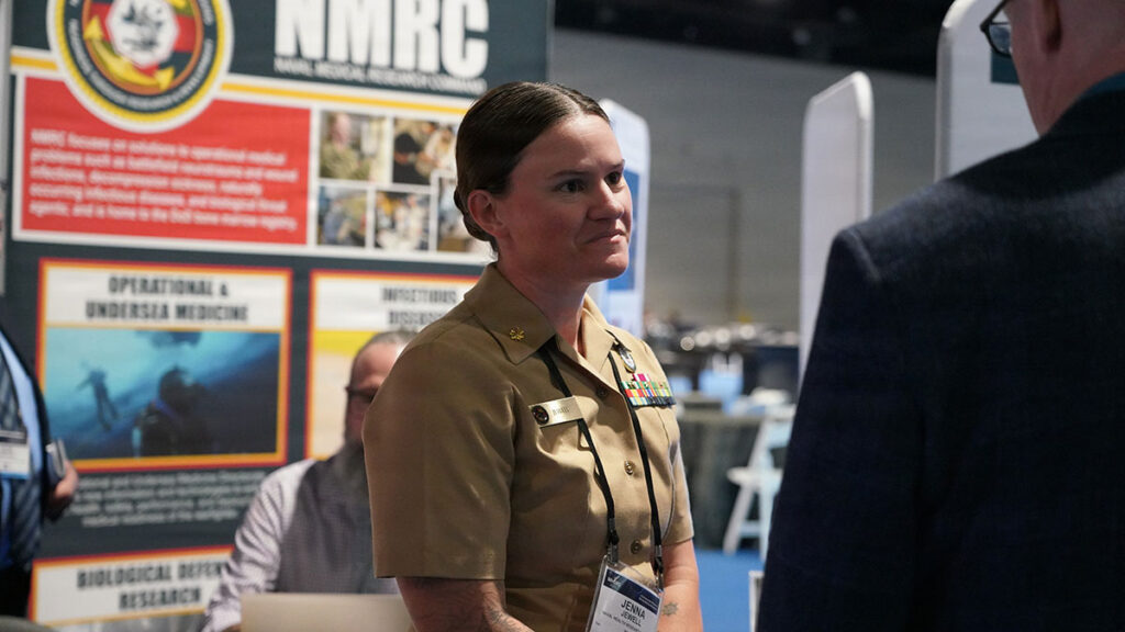 A military officer in uniform stands at a research symposium presentation, engaged in discussion with attendees. Research posters and exhibition displays are visible in the background, showing various research topics and institutional logos. The scene depicts scientific collaboration and knowledge sharing at a major defense research conference.