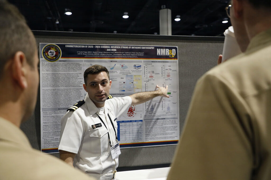 A military researcher in uniform stands at a research poster presentation, gesturing toward the scientific content displayed on the poster board. Attendees view the presentation from the foreground. The poster displays scientific data, charts, graphs, and research methodology. The symposium setting includes professional conference infrastructure and institutional branding visible in the background.