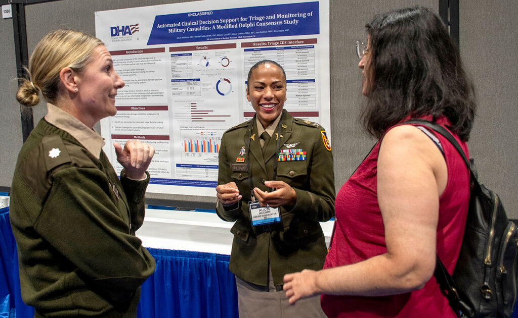 Three military medical professionals engage in discussion at a research poster presentation. The central figure in uniform speaks while gesturing to explain research findings, while two colleagues listen and participate in the conversation. A research poster displaying clinical data, charts, and methodology is displayed behind them. The symposium setting shows professional conference infrastructure and institutional branding.